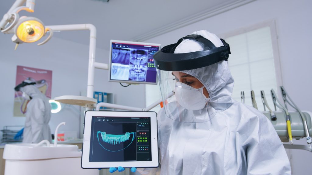 Patient point of view of dentist in protective coverall explaining dental implant treatment using a digital X-ray on a tablet.
